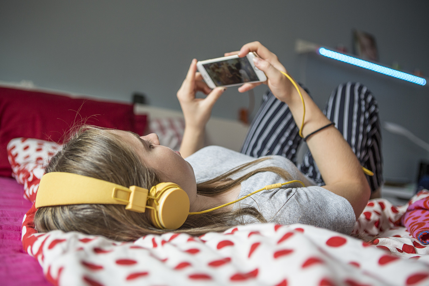 Une adolescente allongée sur un lit qui regarde quelque chose avec des écouteurs sur son smartphone.