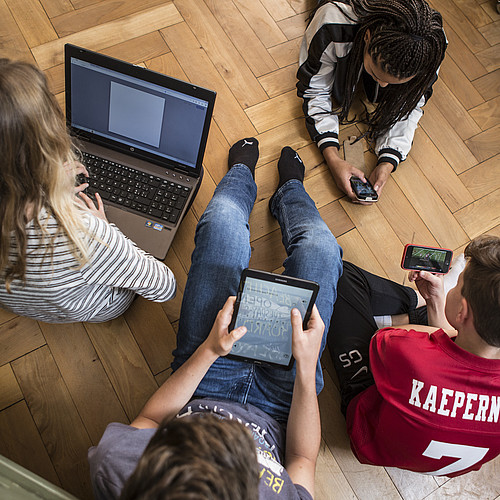 4 adolescents assis ou allongés sur le sol, tous sur un ordinateur portable, une tablette ou un smartphone.