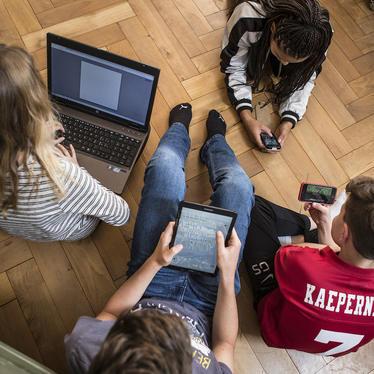 4 adolescents assis ou allongés sur le sol, tous sur un ordinateur portable, une tablette ou un smartphone.