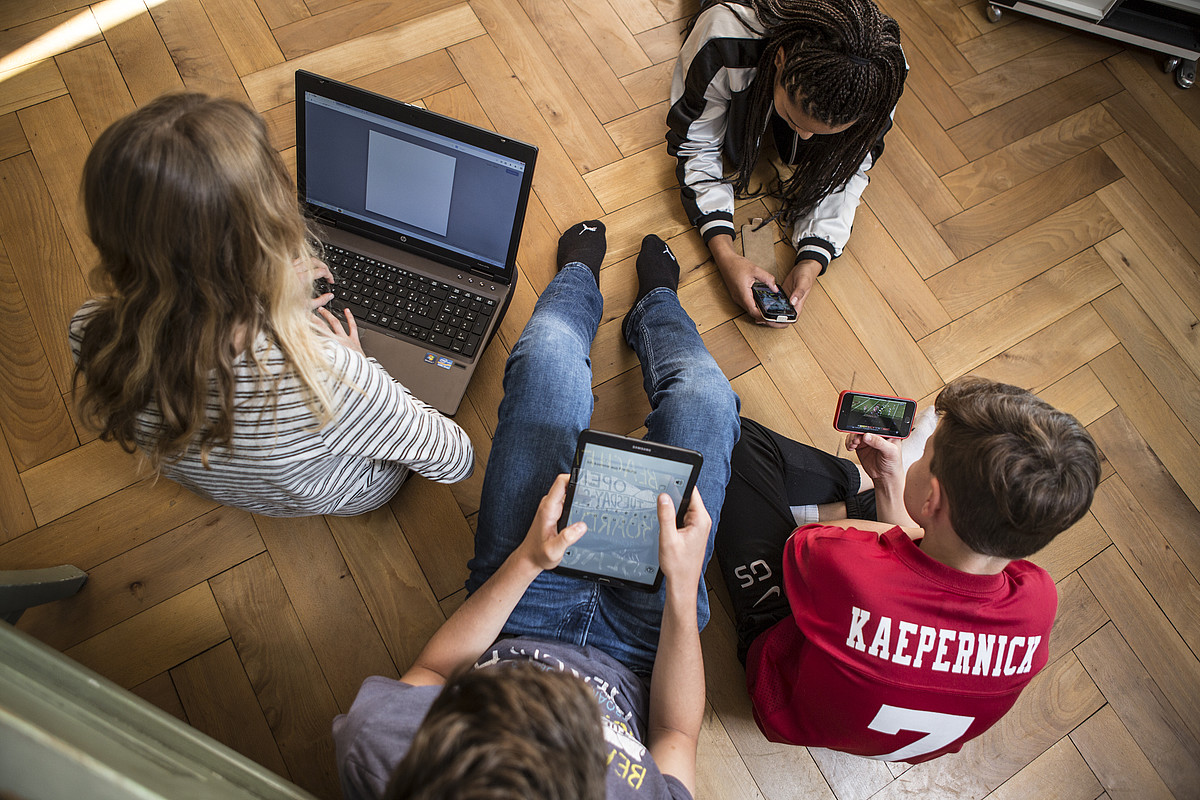 4 adolescents assis ou allongés sur le sol, tous sur un ordinateur portable, une tablette ou un smartphone.