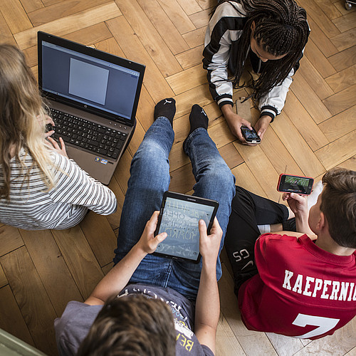 4 adolescents assis ou allongés sur le sol, tous sur un ordinateur portable, une tablette ou un smartphone.