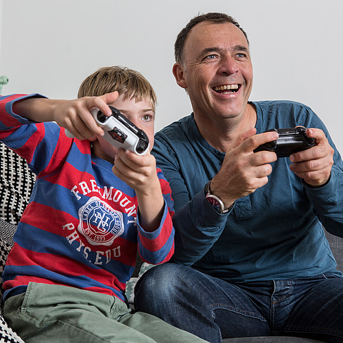 Un père assis sur le canapé avec son fils jouant à des jeux vidéo. 