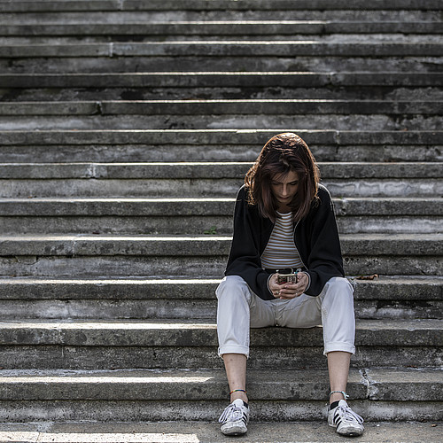 Une jeune femme assise seule dans un escalier consulte son smartphone
