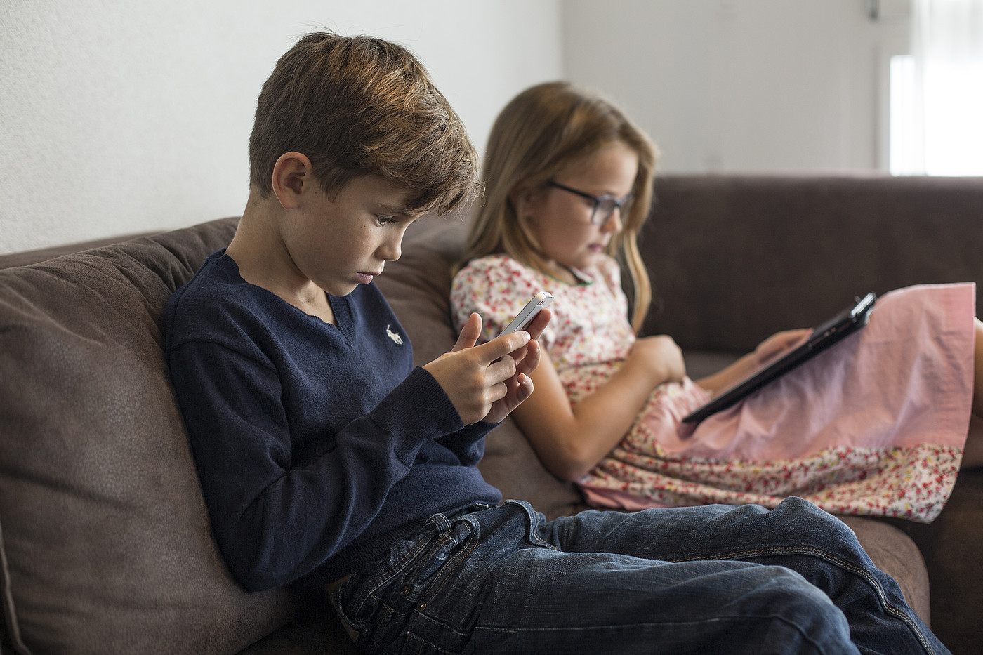 Deux enfants assis sur un canapé avec un smartphone ou une tablette à la main.