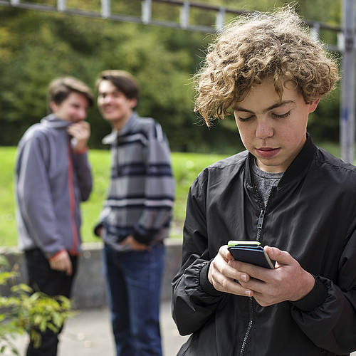 Un adolescent est dehors sur son téléphone portable. En arrière-plan, deux autres jeunes se moquent de lui.