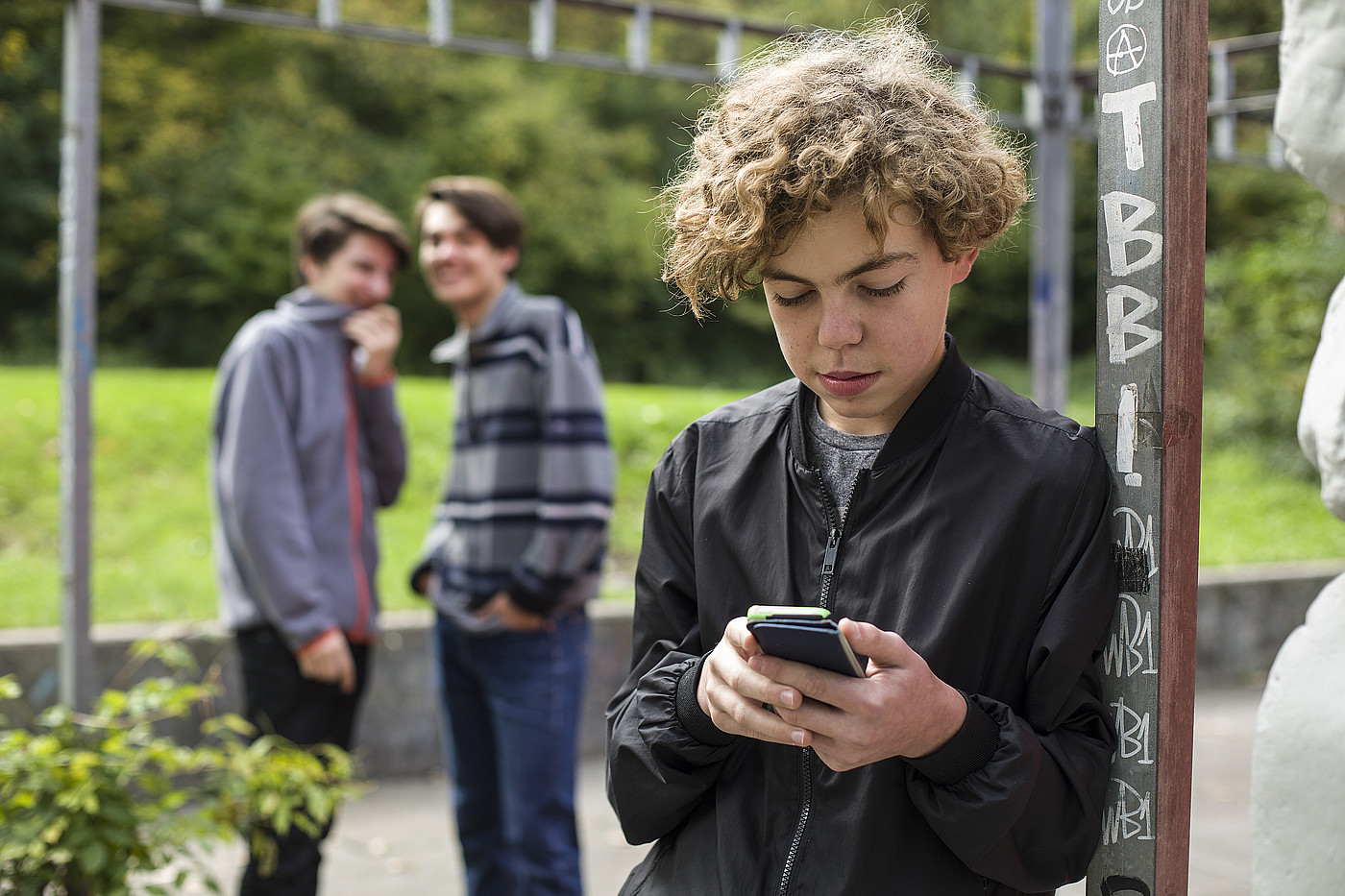 Un adolescent est dehors sur son téléphone portable. En arrière-plan, deux autres jeunes se moquent de lui.