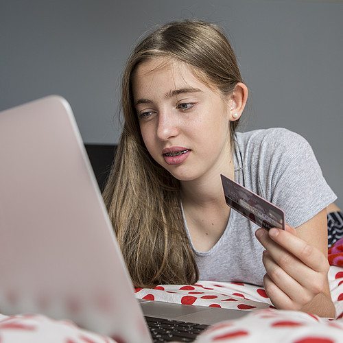 [Translate to Deutsch:] Eine Jugendliche liegt mit dem Laptop auf dem Bett und kauft sich etwas mit der Kreditkarte.