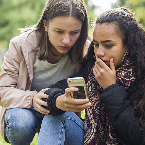 Deux adolescents à l'extérieur regardent un téléphone portable d'un air choqué.