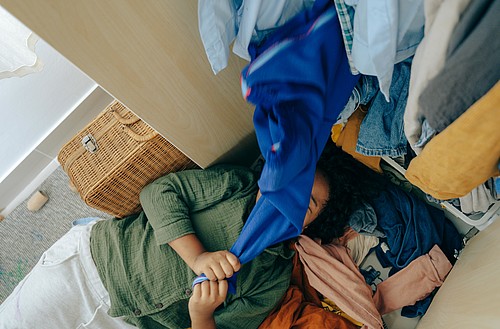 [Translate to Französisch:] Frau liegt vor einem Schrank unter Kleidern begraben.