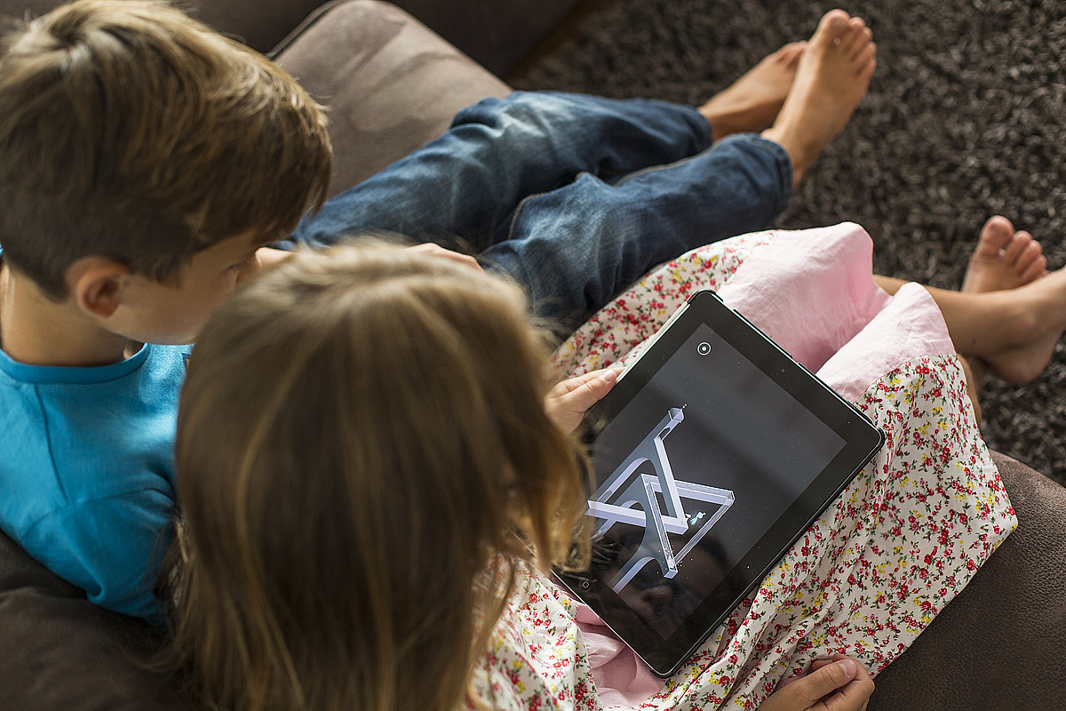 Due bambini seduti su un divano che guardano un tablet.